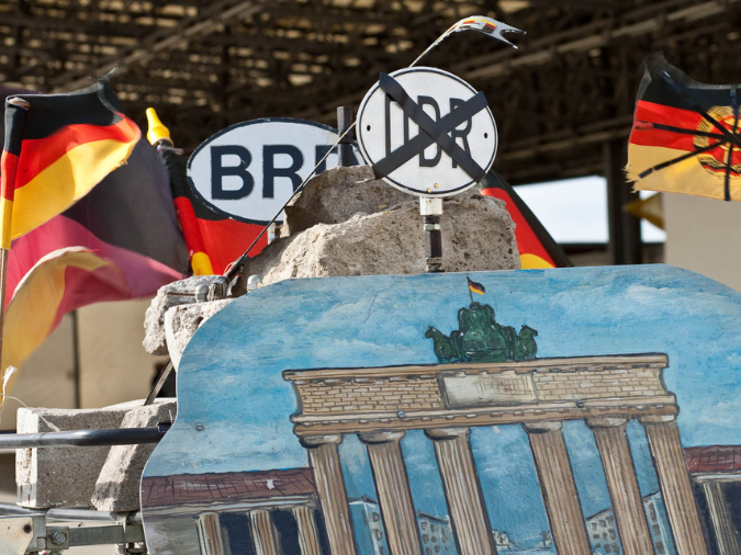 BRD-Flaggen im Hintergrund, eine durchgestrichene DDR-Flagge und ein durchgestrichenes DDR-Schild, im Hintergrund ein BRD-Schild, Steine der Berliner Mauer, ein Gemälde des Brandenburger Tors im Vordergrund