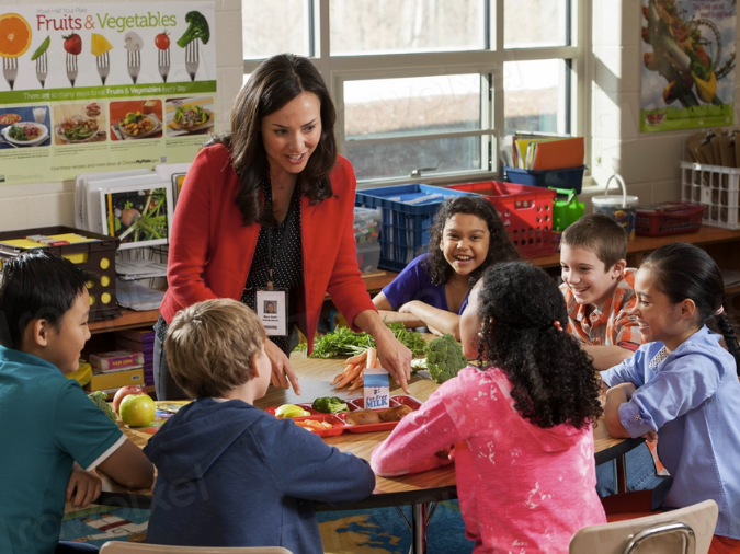 Sechs Kinder sitzen um einen Tisch, darauf liegen gesunde Lebensmittel, eine Lehrerin erklärt ihnen dazu etwas