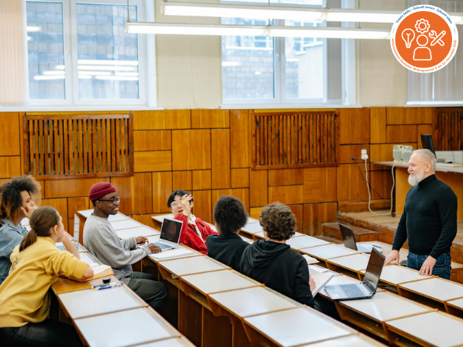 Blick in einen Hörsaal, Dozent steht vor der ersten Tischreihe, sechs Studierende sitzen auf drei Reihen verteilt, drei Studierende haben Laptops vor sich