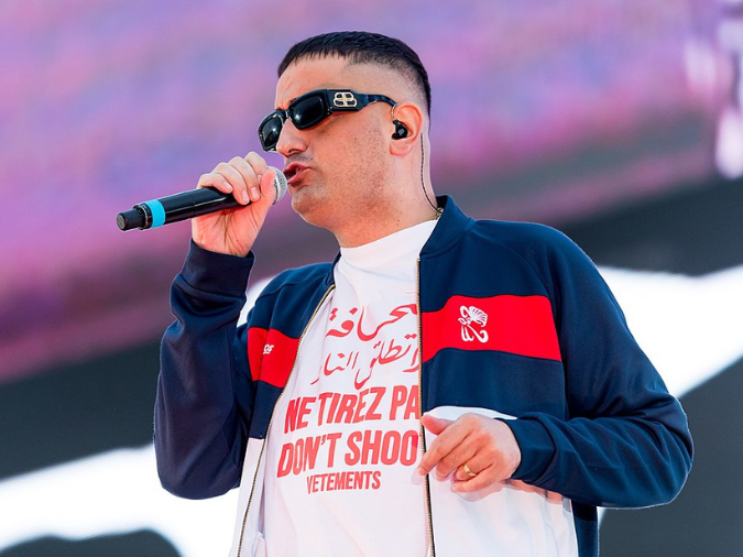 Oberkörperanschnitt-Foto des Rappers Haftbefehl. Er hält ein Mikrofon an seinen Mund, trägt eine blau-weiß-rote Sweatshirt-Jacke und eine schwarze Sonnenbrille.