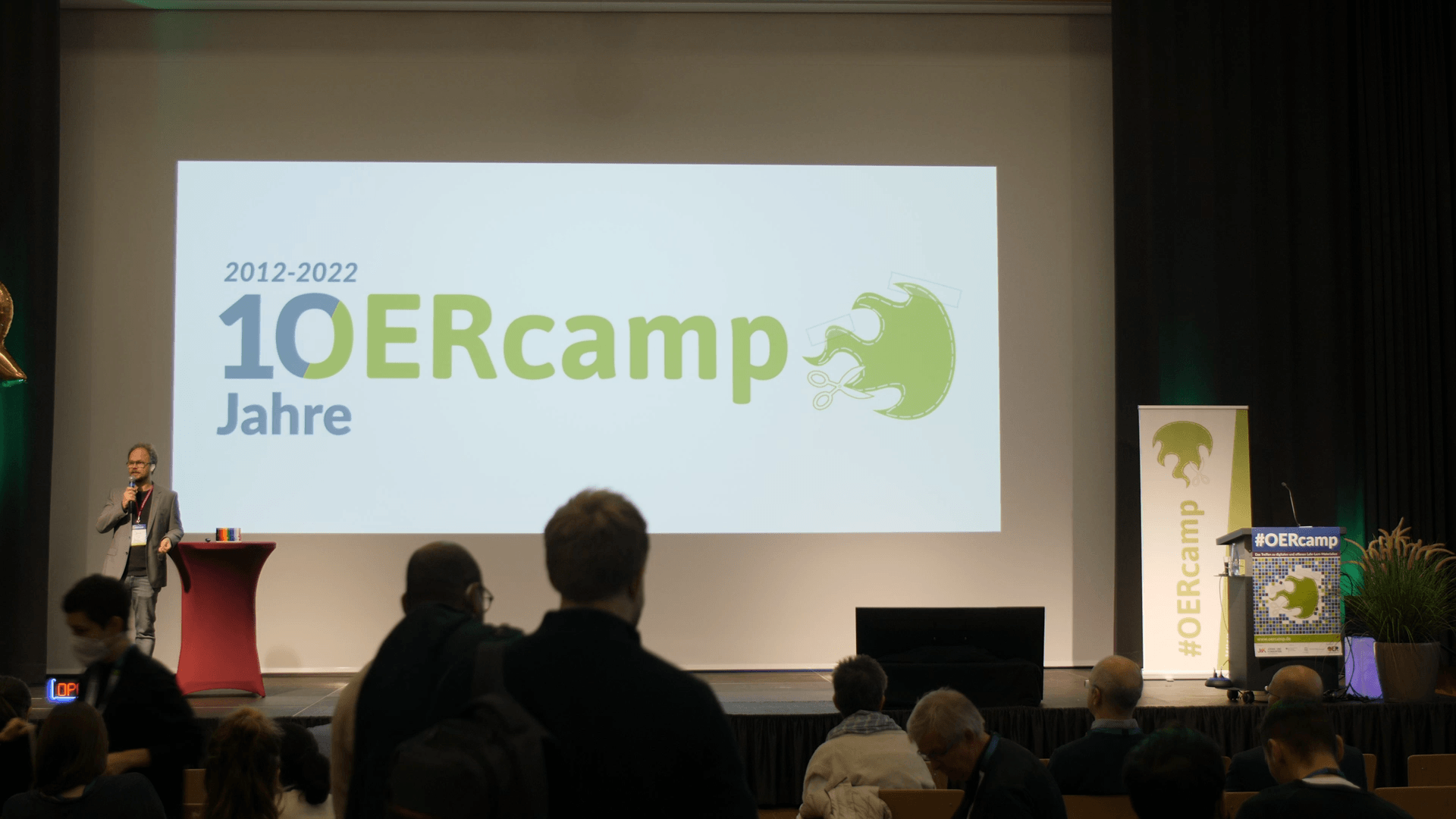 OERcamp2022: Grußworte des BMBF und der DUK, Publikum vor großer Leinwand mit dem Logo 10 Jahre OERcamp und Moderator Jöran Muß-Meerholz auf dem Podium